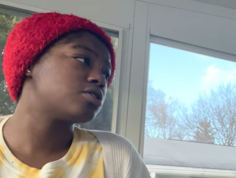 Noelle wears a red fuzzy beanie as she looks to the right and speaks to someone off screen. 